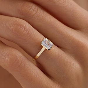 Könnte beinhalten: Ein Goldring mit einem einzelnen, im Smaragdschliff geschliffenen, klaren Edelstein in einer glatten Zargenfassung. Die Ringschiene ist schmal und poliert und unterstreicht durch ihr schlichtes Design die rechteckige Form des Steins.