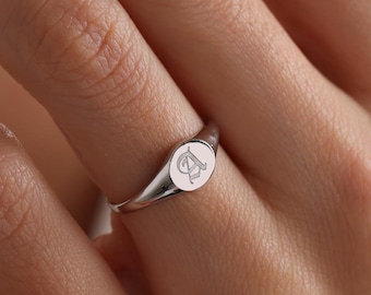 May include: A silver-toned signet ring featuring a polished, smooth band and a circular face engraved with the letter A in a decorative, gothic-style font. The ring has a reflective metallic finish and a simple, clean design on the finger.