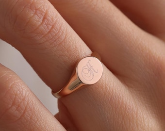 May include: A rose gold signet ring featuring a smooth, polished finish and a circular face. The center of the ring face is engraved with a delicate, cursive letter H, showcasing a clean and simple design that wraps around the finger.