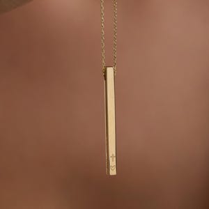 May include: A gold-colored bar pendant necklace. The pendant is rectangular and hangs from a delicate chain. The bar has a small cross and heart engraved on it. The necklace is held up by a person's hand.