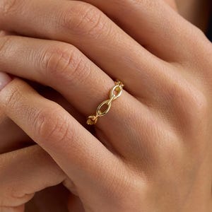 May include: A gold-colored chain link ring. The ring is worn on a finger, with the hands in the foreground. The ring's design features a series of interconnected oval links, creating a continuous chain pattern. The ring is a simple yet elegant piece of jewelry.