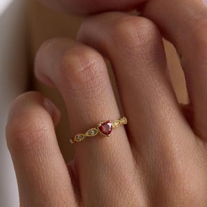 May include: A gold ring featuring a heart-shaped red gemstone. The ring has a detailed design with small, clear gemstones and a textured band. The ring is worn on a finger.