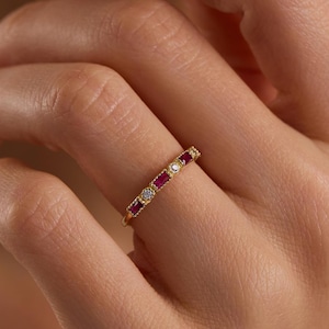 May include: A gold ring featuring alternating red and clear gemstones. The ring is worn on a finger, with the hand and part of a person's face visible. The ring is a delicate piece of jewellery, suitable for everyday wear or special occasions.