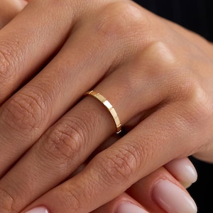 May include: A gold ring with a geometric design. The ring is a thin band with a series of small, rectangular cutouts. The ring is worn on a finger. The background is a dark, solid color.
