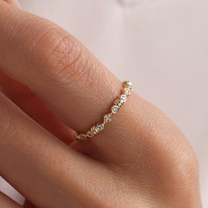 May include: A delicate gold ring featuring a repeating pattern of small, clear gemstones. The ring is worn on a finger, with a white blazer in the background. The ring's design is simple and elegant.