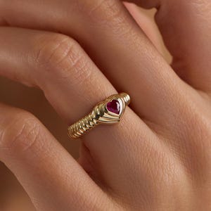 May include: A gold ring with a heart-shaped, red gemstone. The ring has a textured band and is worn on a finger. The ring is a piece of jewelry.