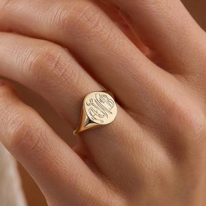 Puede incluir: Un anillo de sello de oro con acabado pulido y una cara circular grabada con un monograma estilizado e intrincado. El anillo tiene una banda lisa y cónica que se asienta sobre el dedo, mostrando un diseño clásico y refinado.