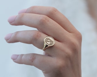May include: A gold signet ring with a round, engraved design. The ring is worn on the finger of a person's hand.