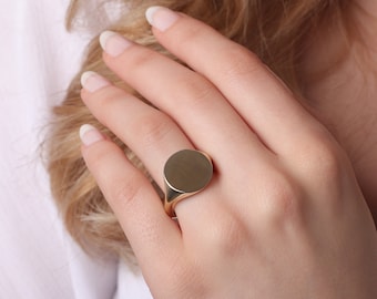 May include: A polished gold signet ring featuring a smooth, flat, circular face. The ring has a solid, minimalist band design with a reflective metallic finish that catches the light, showcasing a clean and simple aesthetic for everyday wear.