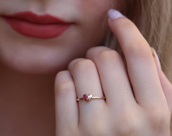 May include: A gold ring with a heart-shaped, red gemstone. The ring has a slender band and a heart-shaped stone at the centre. The ring is worn on a finger, highlighting its simple design. Jewellery.