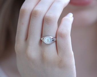 May include: A silver ring with a white oval stone surrounded by smaller stones. The ring is on a person's finger.