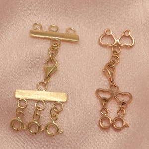 May include: Two gold-toned jewelry extenders. The extender on the left has a bar with three loops and a dangling chain with three loops. The extender on the right has two heart-shaped loops and a dangling chain with two heart-shaped loops.