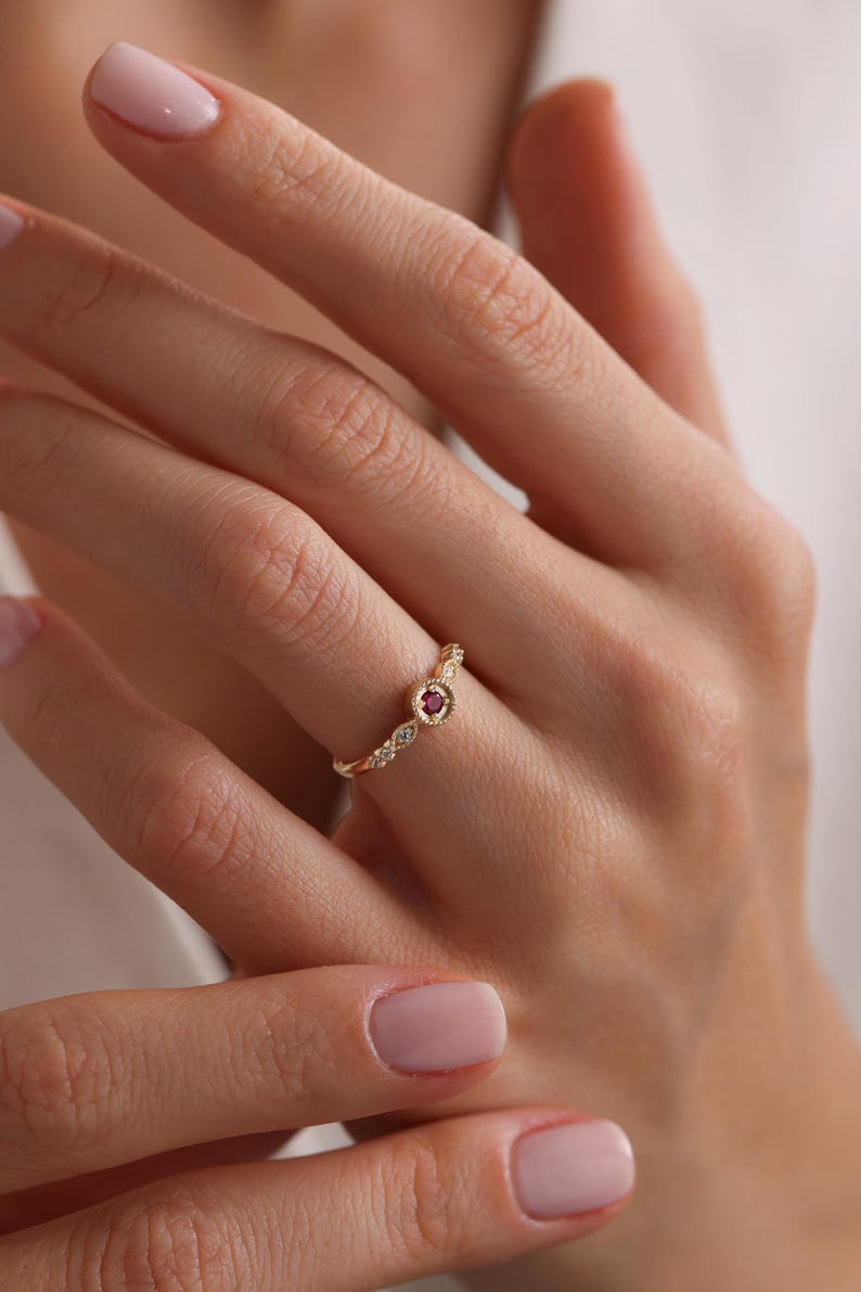 May include: A gold ring with a small red gemstone and smaller white gemstones set in a delicate, curved design.