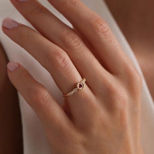 May include: A delicate gold ring featuring a small red gemstone surrounded by tiny diamonds. The ring is worn on a finger with a light pink nail polish.