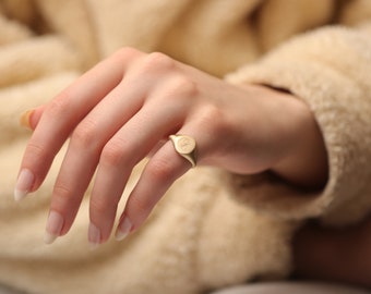 May include: A gold signet ring with a rounded oval top. The ring is worn on the ring finger of a hand.