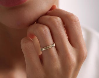 May include: A gold band ring featuring a repeating vertical ribbed or fluted texture around the entire circumference. The ring has a polished metallic finish and a consistent width, showcasing a structured design with clean, parallel lines.