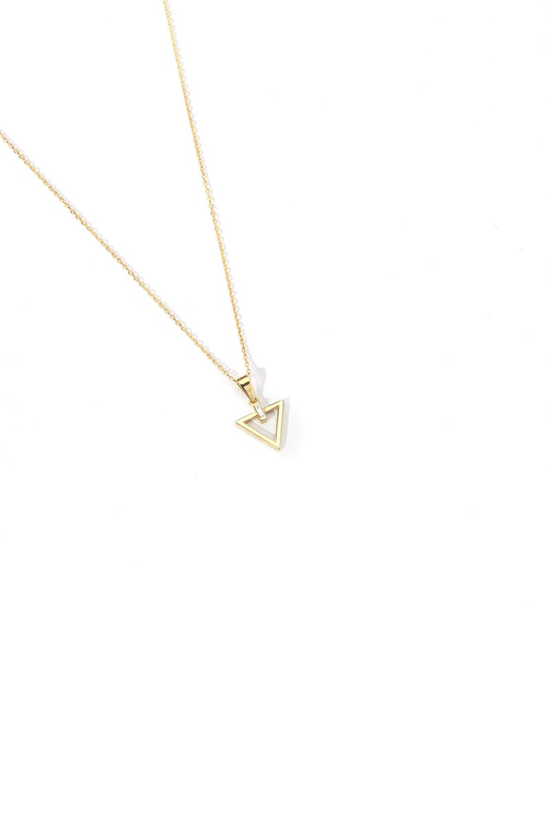 Puede incluir: Un delicado collar dorado con un colgante triangular. El colgante tiene un dise&ntilde;o geom&eacute;trico con una peque&ntilde;a piedra transparente en el centro. La cadena es fina y elegante, adecuada para el uso diario.