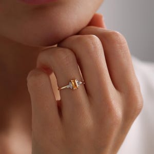 Op de afbeelding: Een gouden ring met een centrale rechthoekige gele edelsteen in smaragdgeslepen vorm, geflankeerd door twee kleine heldere baguette-geslepen stenen aan elke kant. De ringband is dun en gemaakt van gepolijst goud met een klassiek ontwerp.