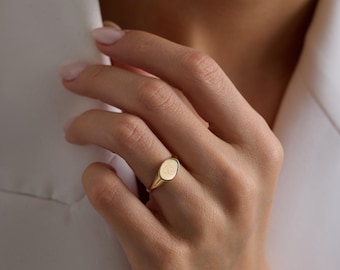 May include: A polished gold signet ring featuring a smooth, oval-shaped face with a subtle, engraved monogram detail. The band is solid and tapers slightly toward the back, showcasing a clean, reflective metallic finish throughout the design.