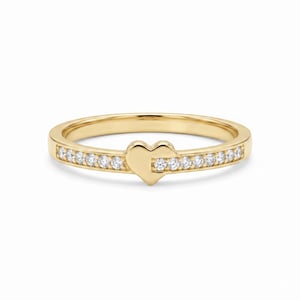 May include: A gold ring featuring a central heart-shaped motif flanked by a row of small, clear, round-cut stones embedded along the band on both sides. The metal has a polished, reflective finish, and the design is symmetrical and slender.
