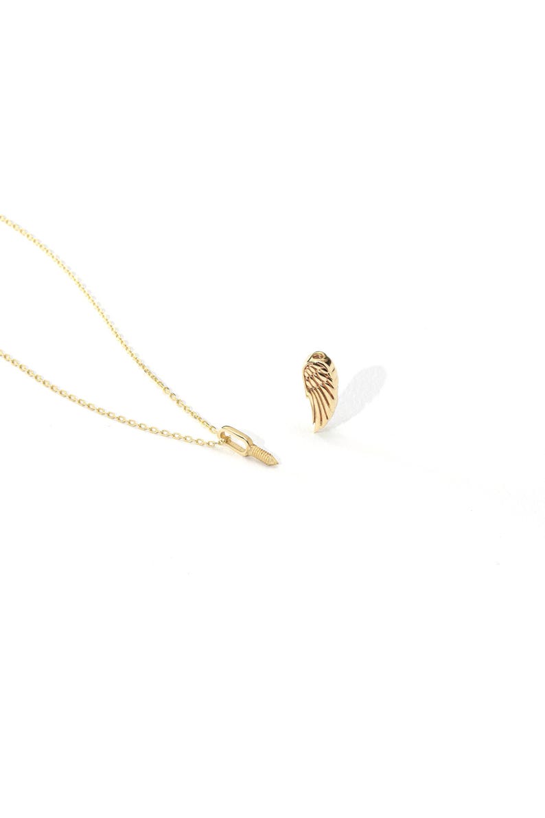 May include: A gold-colored necklace with a small pendant and a matching wing-shaped earring. The necklace chain is delicate, and the pendant has a textured design. The earring is a detailed wing shape, casting a shadow on the white background.