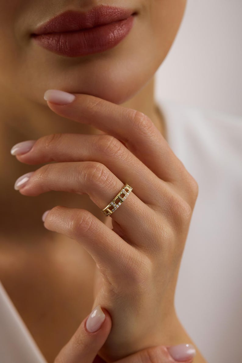 May include: A gold ring with a repeating rectangular design, each section adorned with small, clear gemstones. The ring is worn on a finger, showcasing its intricate details and sparkling appearance. The ring is a piece of jewelry.