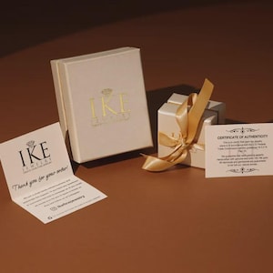 a jewelry box with a gold ribbon, a small white box with a gold ribbon, and a white card with text on a brown background.