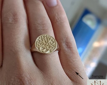 May include: A gold signet ring with a round, raised design featuring Arabic calligraphy. The ring is worn on a person's finger.