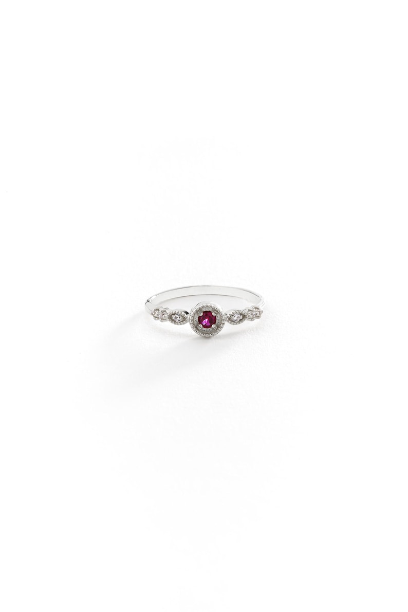 May include: A silver ring featuring a red gemstone set within a halo of smaller white gemstones. The ring has a delicate, vintage-inspired design.