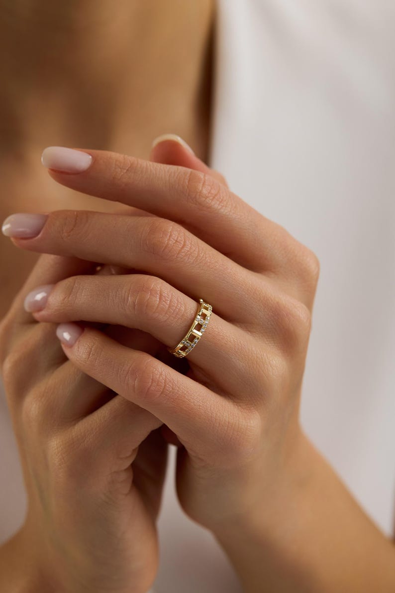 Puede incluir: Un anillo dorado con un dise&ntilde;o de eslabones de cadena, engastado con peque&ntilde;as piedras preciosas transparentes. El anillo se lleva en un dedo, mostrando sus intrincados detalles y acentos brillantes. El dise&ntilde;o del anillo es moderno y elegante.