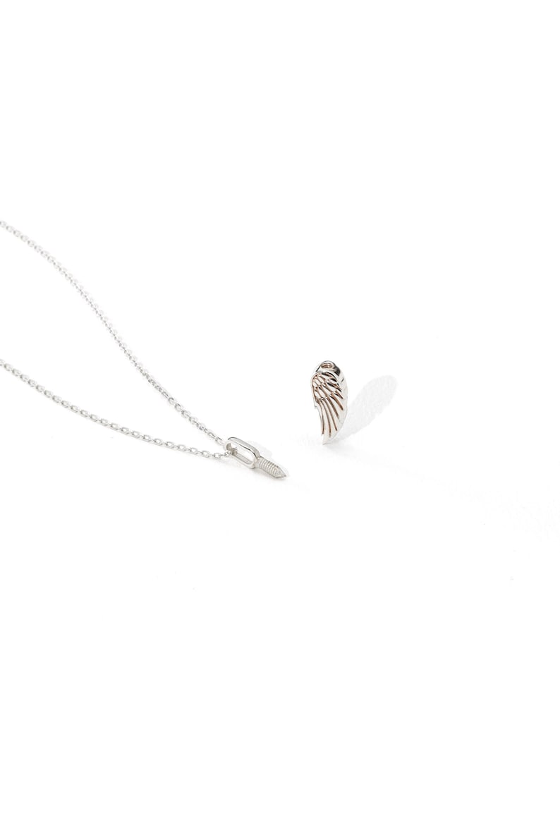 May include: A silver necklace with a delicate chain and a small pendant. A matching silver wing-shaped earring is also visible, casting a shadow on the white background. The jewelry is likely made of sterling silver.