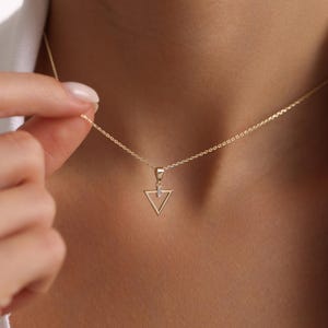 Puede incluir: Un delicado collar de oro con un colgante triangular geom&eacute;trico con un peque&ntilde;o acento brillante. El colgante cuelga de una fina cadena, creando un aspecto minimalista y elegante. El collar se muestra sobre un fondo de tono de piel.
