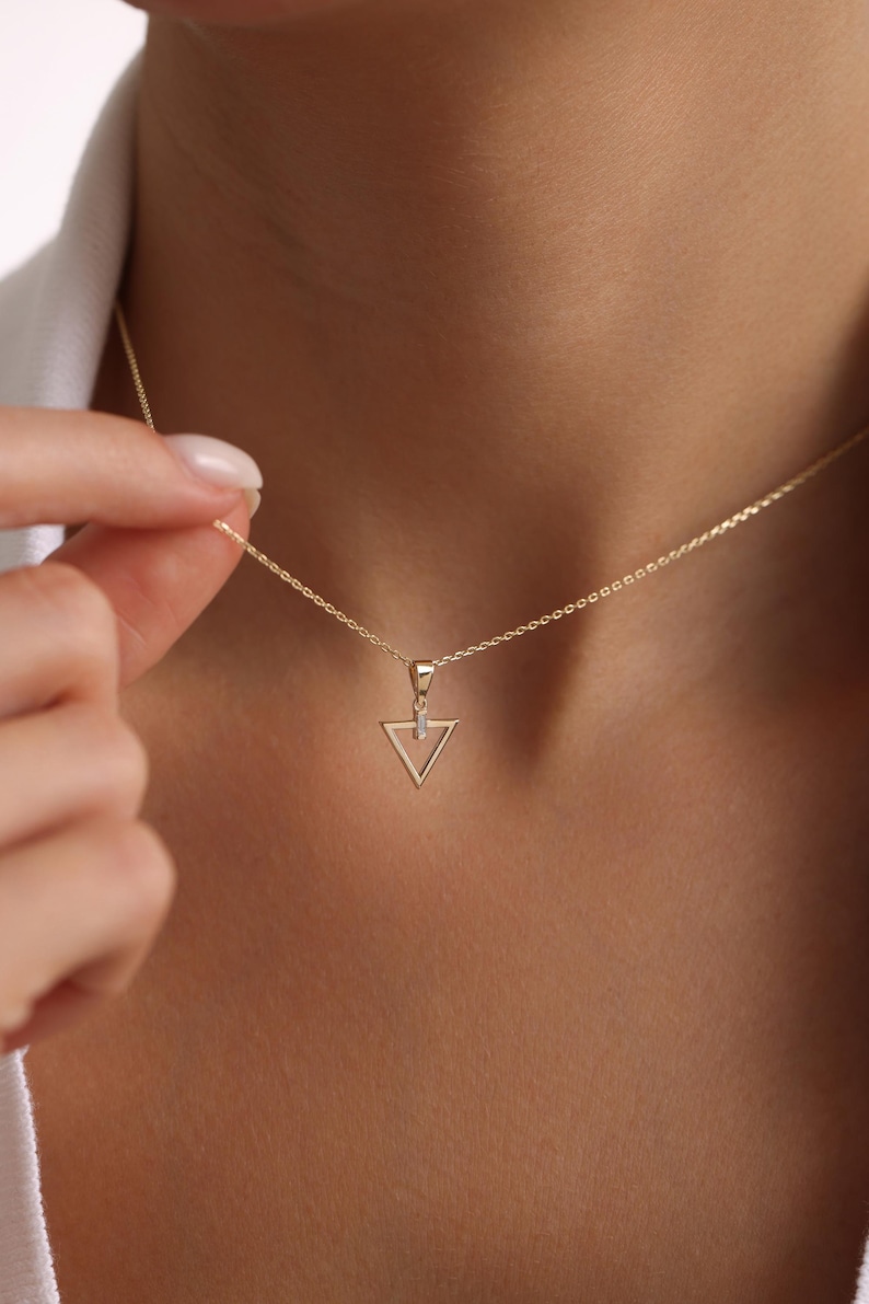 Puede incluir: Un delicado collar de oro con un colgante triangular geom&eacute;trico con un peque&ntilde;o acento brillante. El colgante cuelga de una fina cadena, creando un aspecto minimalista y elegante. El collar se muestra sobre un fondo de tono de piel.