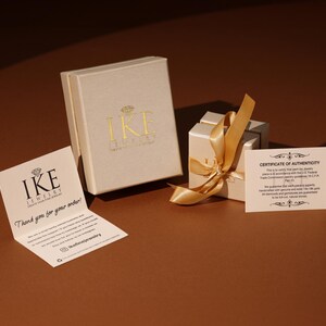 Puede incluir: Una presentaci&oacute;n de joyas con dos cajas beige, una abierta con una cinta dorada y una caja cerrada con letras doradas. Las tarjetas adjuntas incluyen una nota de agradecimiento y un certificado de autenticidad, todo sobre una superficie marr&oacute;n.