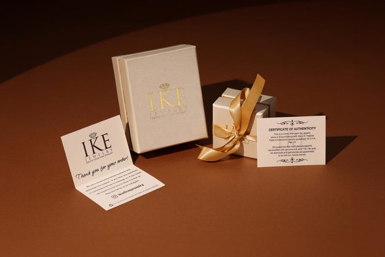 Puede incluir: Una presentaci&oacute;n de joyas con dos cajas beige, una abierta con una cinta dorada y una caja cerrada con letras doradas. Las tarjetas adjuntas incluyen una nota de agradecimiento y un certificado de autenticidad, todo sobre una superficie marr&oacute;n.