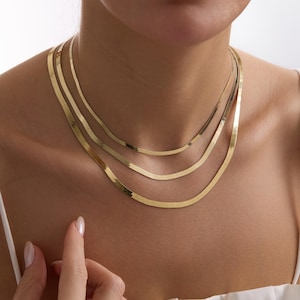 Könnte beinhalten: Ein Set aus drei geschichteten goldenen Fischgrätenketten. Jede Halskette zeichnet sich durch ein flaches, flexibles und poliertes Metallglied-Design aus, das eng am Hals in einer abgestuften Anordnung liegt und einen gestapelten Look erzeugt.