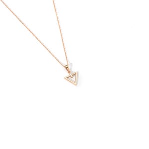 Puede incluir: Un delicado collar dorado con un colgante triangular. El colgante presenta un dise&ntilde;o geom&eacute;trico con una peque&ntilde;a piedra transparente en el centro. La cadena es fina y el collar est&aacute; sobre un fondo blanco.
