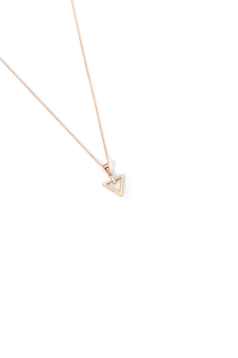 Puede incluir: Un delicado collar dorado con un colgante triangular. El colgante presenta un dise&ntilde;o geom&eacute;trico con una peque&ntilde;a piedra transparente en el centro. La cadena es fina y el collar est&aacute; sobre un fondo blanco.