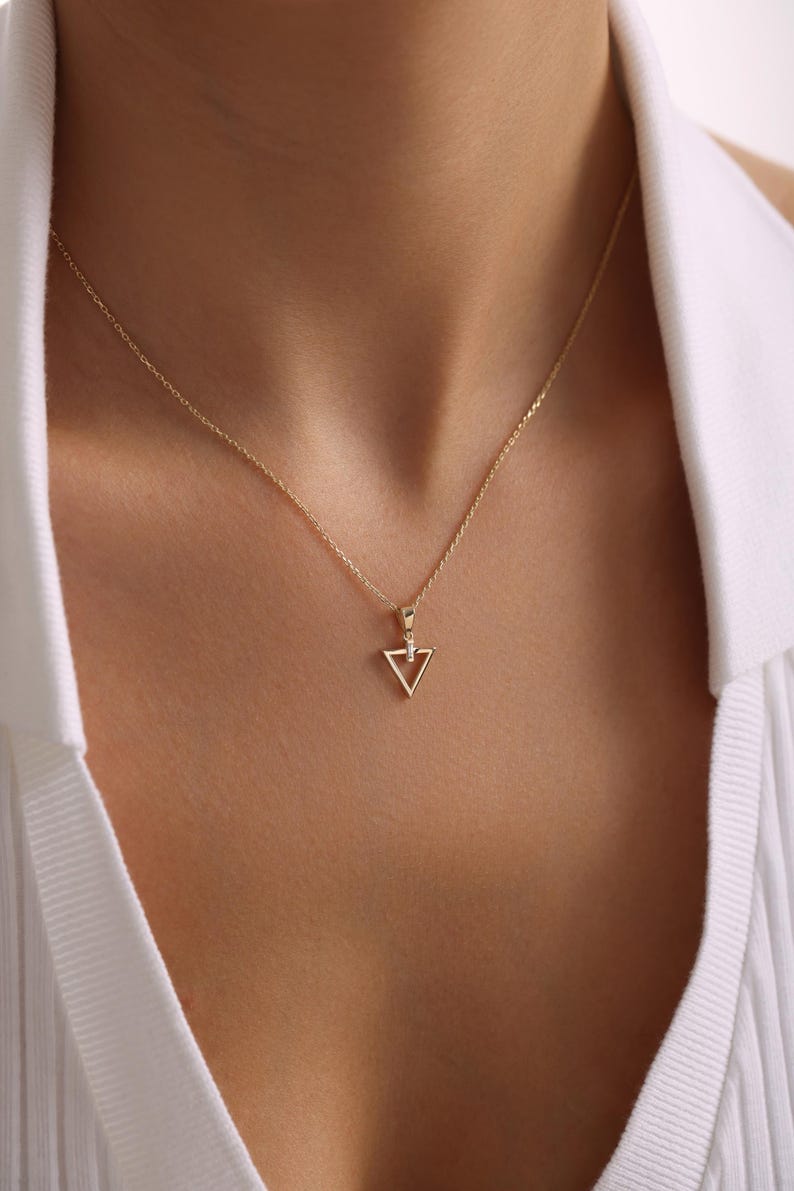 Puede incluir: Un delicado collar de oro con un colgante triangular y un peque&ntilde;o acento brillante. El colgante cuelga de una fina cadena, creando un aspecto minimalista y elegante. El collar se muestra sobre una camisa blanca.