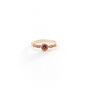 May include: A delicate gold ring featuring a small ruby gemstone surrounded by tiny diamonds. The ring has a vintage-inspired design with intricate details.