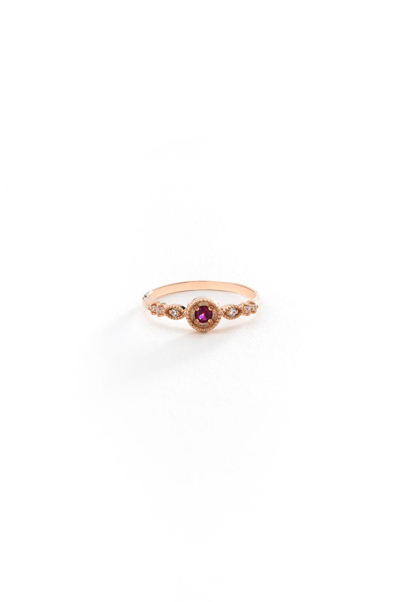 May include: A delicate gold ring featuring a small ruby gemstone surrounded by tiny diamonds. The ring has a vintage-inspired design with intricate details.