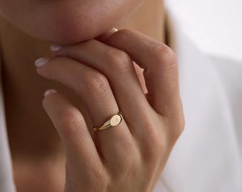 May include: A gold signet ring featuring a smooth, polished band and a small, oval-shaped face. The center of the oval face is engraved with a delicate, circular monogram design. The ring has a warm, metallic yellow finish and a simple, clean profile.
