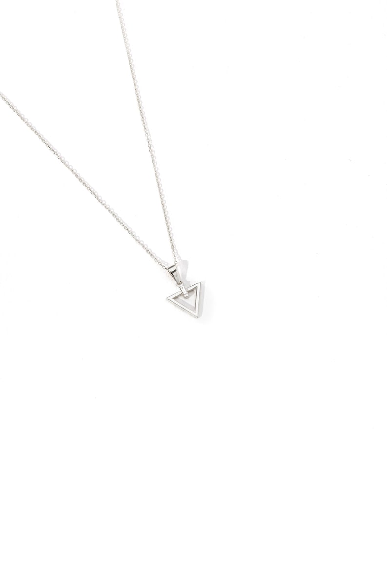 Puede incluir: Collar plateado con un colgante triangular hueco. El colgante cuelga de una cadena fina. El collar se muestra sobre un fondo blanco.