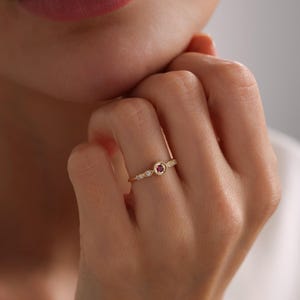 May include: A delicate gold ring featuring a small red gemstone surrounded by tiny white gemstones.