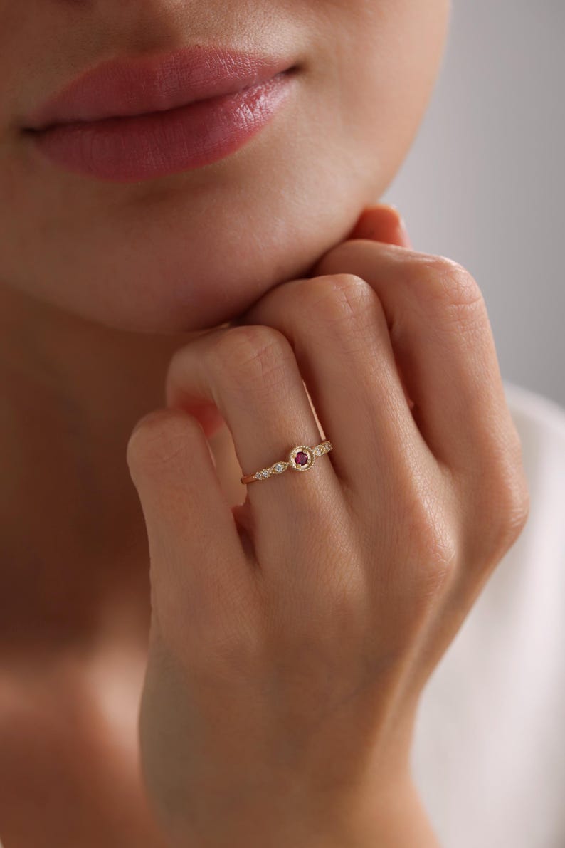 May include: A delicate gold ring featuring a small red gemstone surrounded by tiny white gemstones.