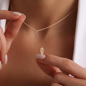 May include: A delicate gold-colored necklace with a small wing pendant. The necklace is held by a person, showcasing the intricate details of the wing charm. The chain is fine and elegant, suitable for everyday wear.