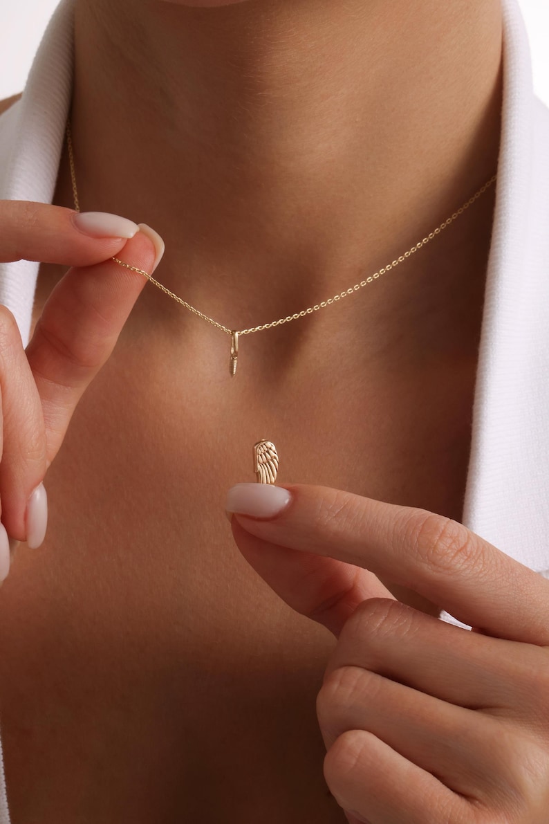May include: A delicate gold-colored necklace with a small wing pendant. The necklace is held by a person, showcasing the intricate details of the wing charm. The chain is fine and elegant, suitable for everyday wear.