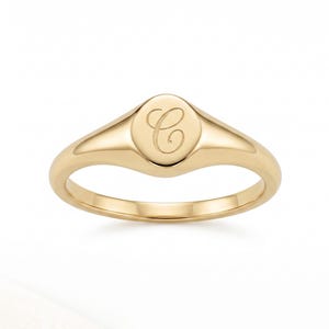 May include: A gold-colored signet ring with a polished finish. The ring features an oval face with the cursive letter "C" engraved in the center. The band tapers towards the back.