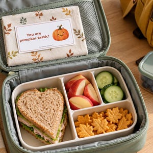 AI-generated lifestyle mockup: a pumpkin encouragement note on a fall napkin inside a sage school lunchbox beside a packed lunch. Listing includes printable PDF notes only; food and lunchbox are not included.