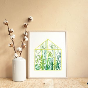 May include: A framed print of a yellow-outlined greenhouse filled with various green plants and ferns. The print is on a white background and is displayed on a light wood surface.